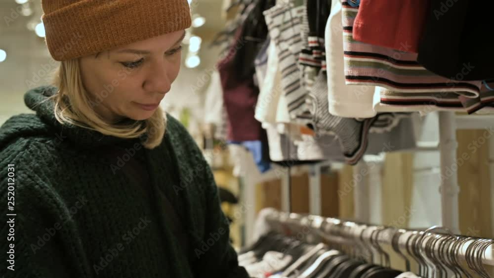 A girl in a green sweater and a yellow hat walks through a store of things and chooses what to buy. Touches things on hangers and looks at price tags A girl in a green sweater and a yellow hat walks through a store of things and chooses what to buy. Touches things on hangers and looks at price tags