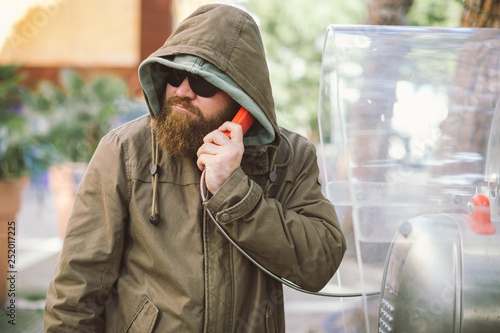Portrait of young bearded man using public phone wearing hoodie (jacket) and black sunglasses - informant, spy, secret agent and fake news concept