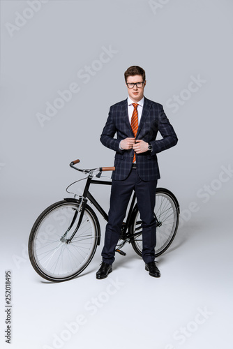 stylish businessman in formal wear posing with bicycle on grey