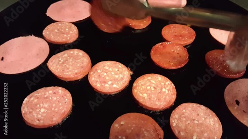 Closeup of a hand with hygienic gloves holding tong and frying Turkish sausage (sucuk) in a black pan 