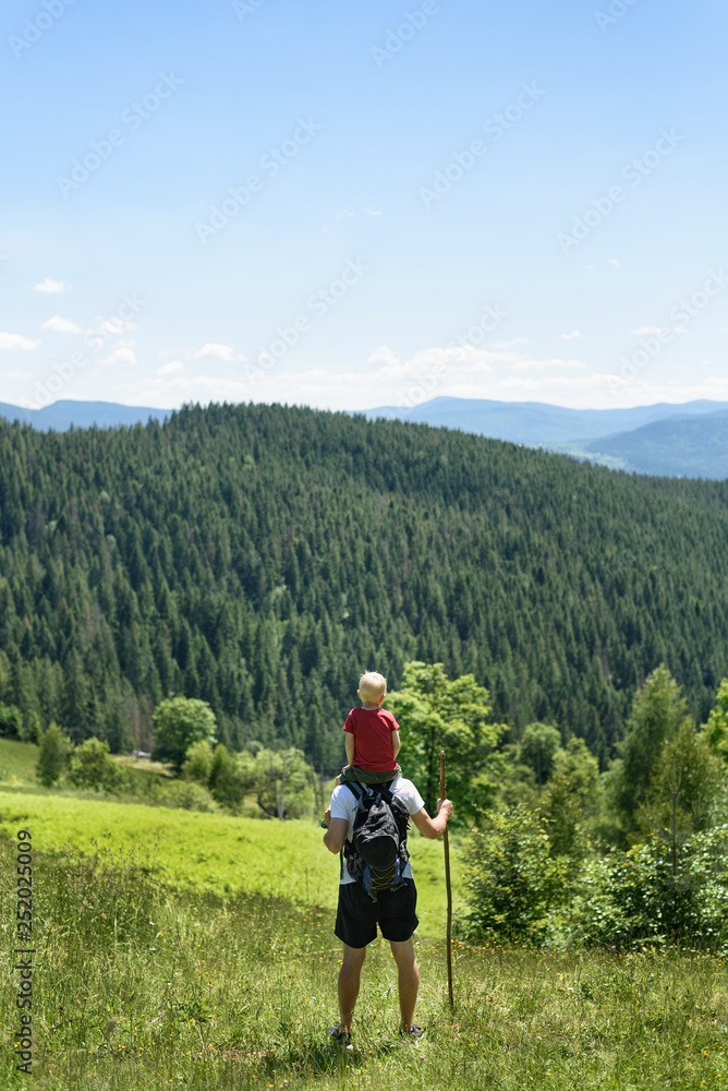 Father with son on his shoulders standing with staff in the background of green forest, mountains and sky with clouds. Back view Father with son on his shoulders standing with staff in the background of green forest, mountains and sky with clouds. Back view