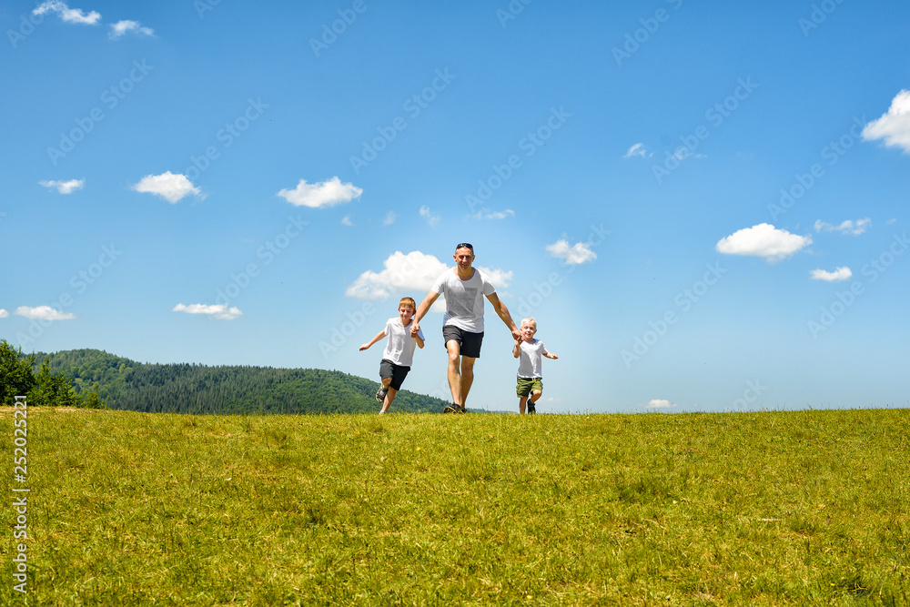 Father with two young children running hand in hand on the green field on a background of blue sky and clouds. Fatherhood and friendship
