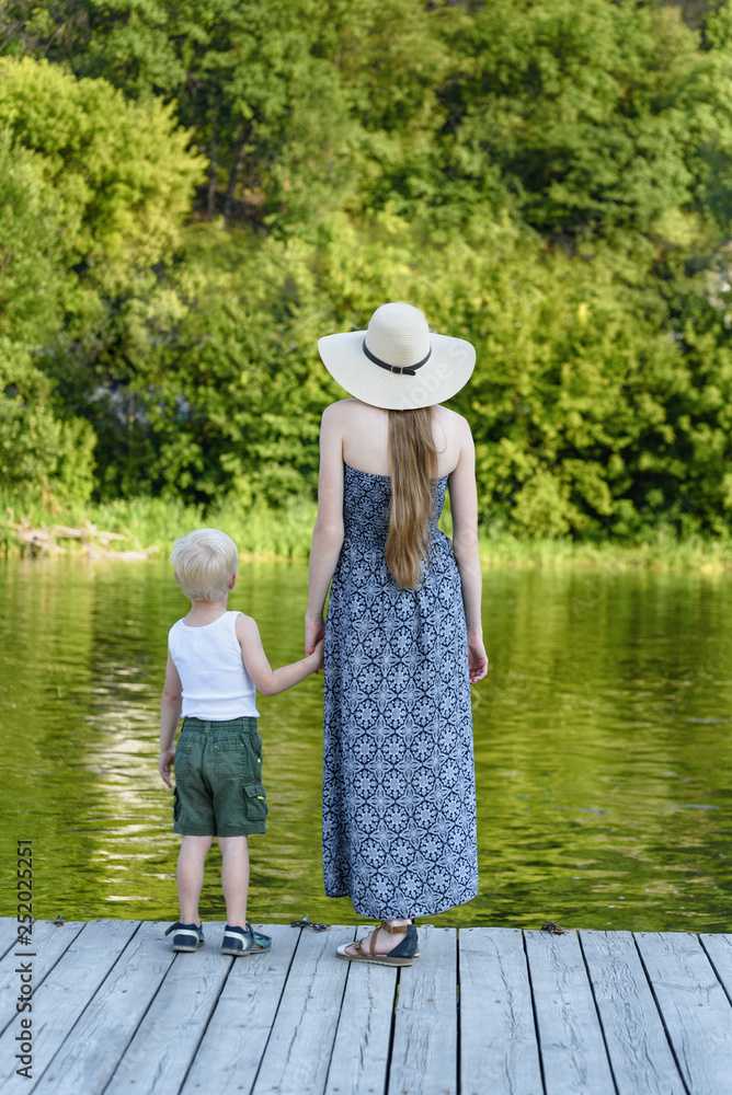 Beautiful mother with her little son are standing on the pier by the river. Back view. Vertical frame Beautiful mother with her little son are standing on the pier by the river. Back view. Vertical frame
