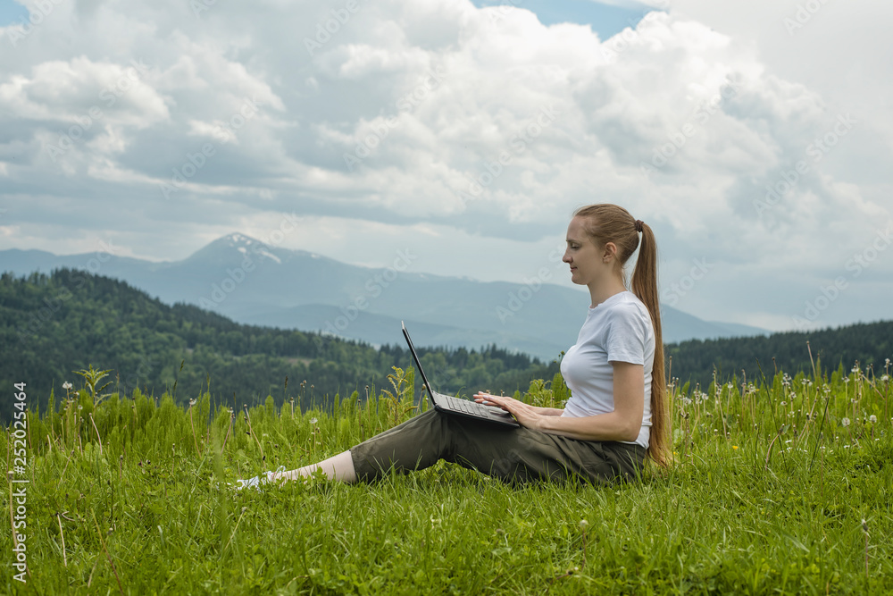 Beautiful girl with a laptop sitting on green grass on a background of mountains