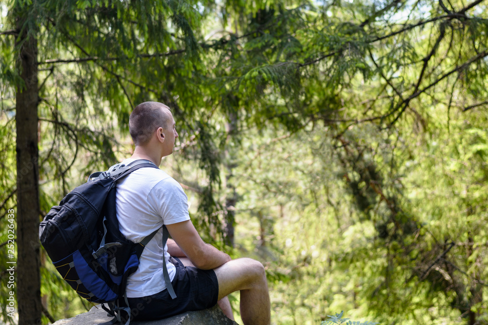 Obraz premium Young man with a backpack sits on a rock in the coniferous forest. Side view