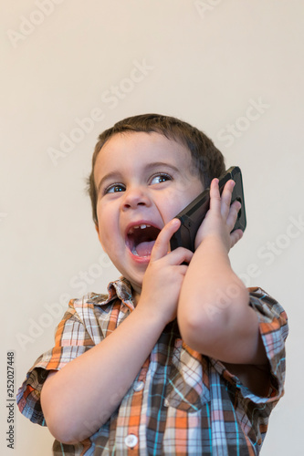 Cute little boy talking by mobile phone on light background. Happy little boy standing and talking on smartphone at home. Technology, education and lifestyle concept. Copy space. vertical photo