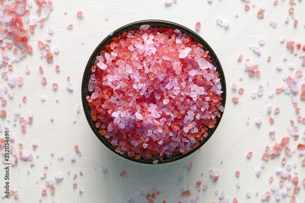 The fragrant and revitalizing sea salt for a bath in a brown bowl stands on a white table in the center, with salt crystals scattered around it.   Close up on a soft natural