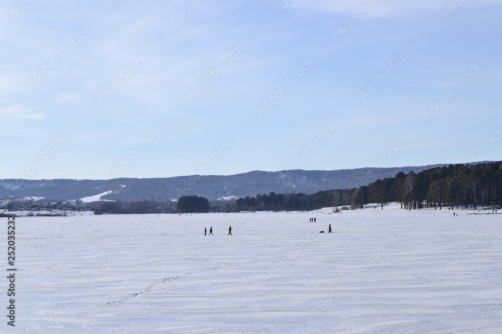 Frozen pond, forest and mountains. Winter landscape. People walk on the snowy surface of the pond. There are mountains in the background.