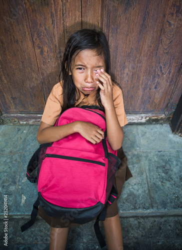 7 or 8 years child in school uniform sitting outdoors on the floor crying sad and depressed holding her backpack suffering bullying and abuse problem feeling alone and helpless