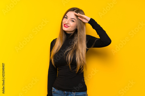 Young pretty woman over yellow background saluting with hand