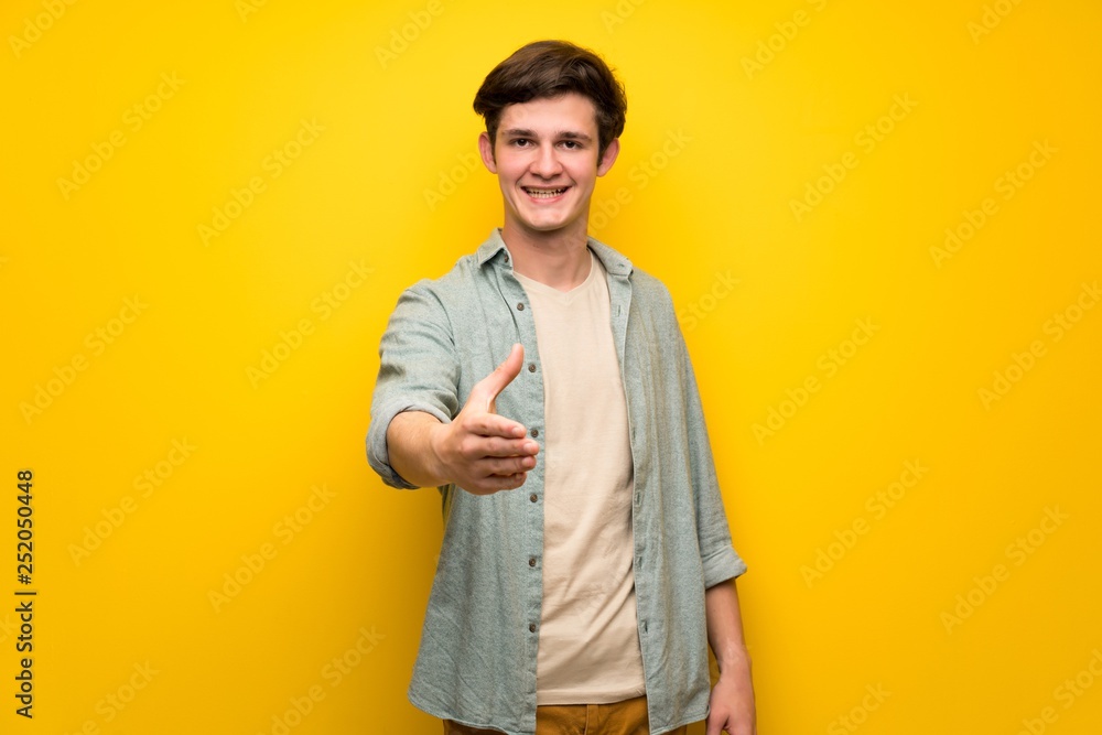 Teenager man over yellow wall shaking hands for closing a good deal Teenager man over yellow wall shaking hands for closing a good deal