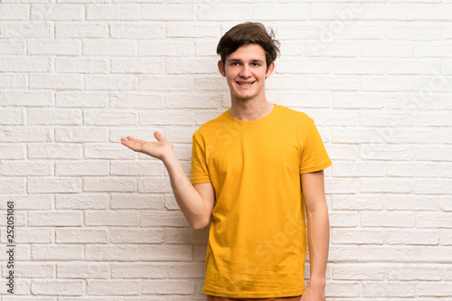Teenager man over white brick wall holding copyspace imaginary on the palm to insert an ad