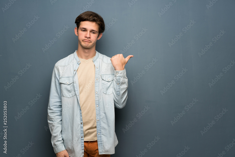 Teenager man with jean jacket over grey wall unhappy and pointing to the side Teenager man with jean jacket over grey wall unhappy and pointing to the side