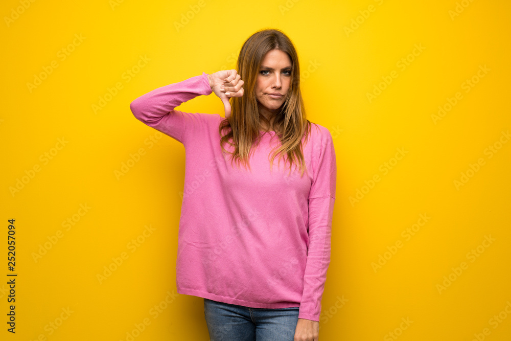 Woman with pink sweater over yellow wall showing thumb down with negative expression Woman with pink sweater over yellow wall showing thumb down with negative expression