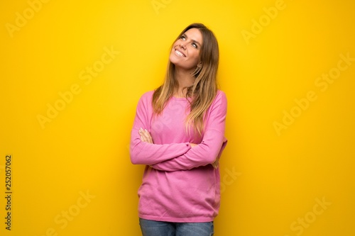 Woman with pink sweater over yellow wall looking up while smiling