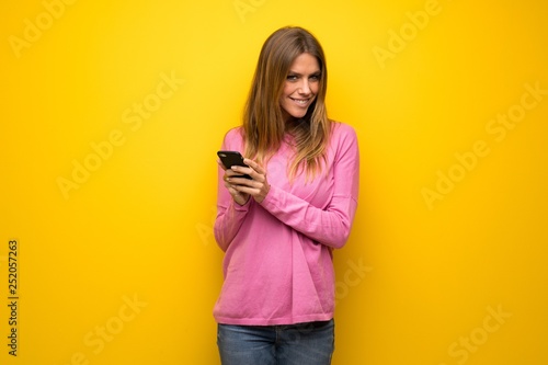 Woman with pink sweater over yellow wall sending a message with the mobile