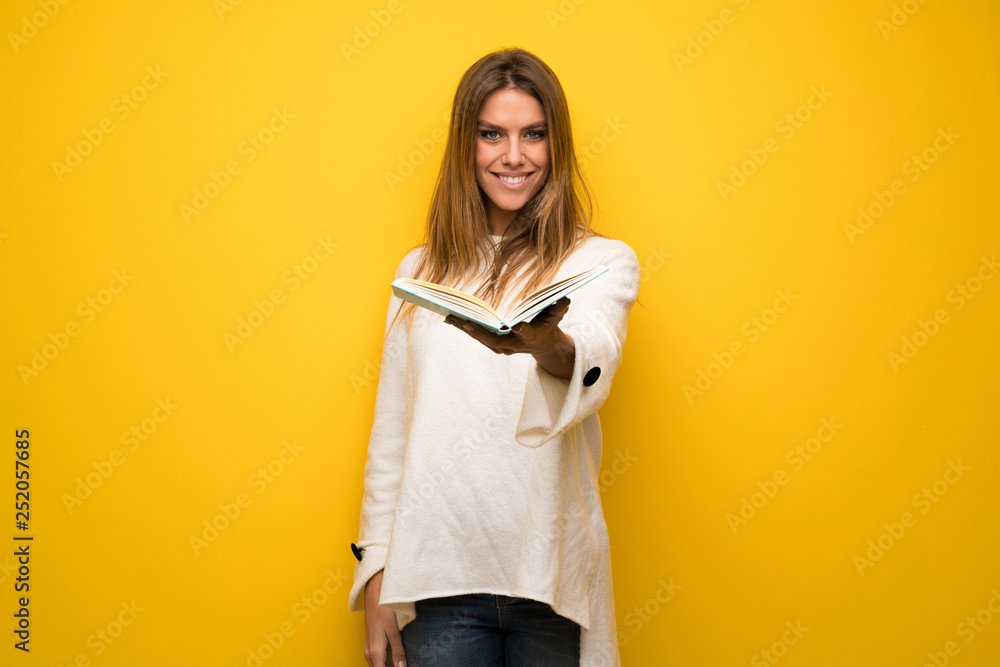 Blonde woman over yellow wall holding a book and giving it to someone Blonde woman over yellow wall holding a book and giving it to someone