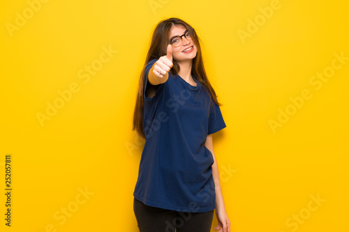 Young woman with glasses over yellow wall giving a thumbs up gesture because something good has happened © luismolinero