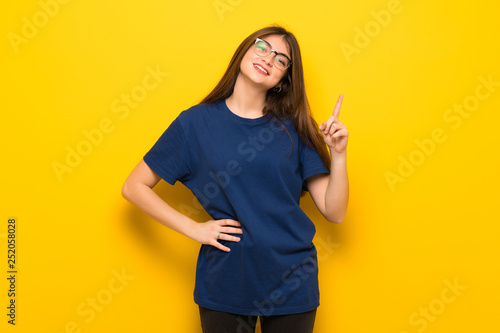Young woman with glasses over yellow wall showing and lifting a finger in sign of the best