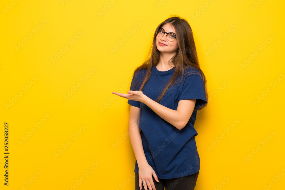 Young woman with glasses over yellow wall presenting an idea while looking smiling towards Young woman with glasses over yellow wall presenting an idea while looking smiling towards