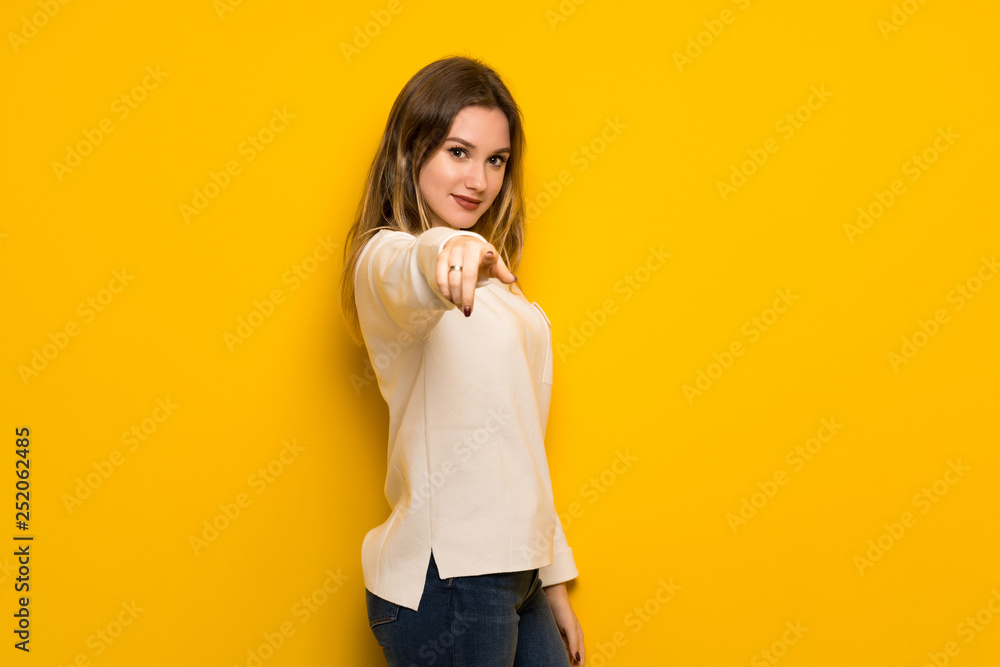Teenager girl over yellow wall points finger at you with a confident expression Teenager girl over yellow wall points finger at you with a confident expression