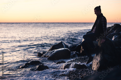 Silhouette of a slim young guy leaning on a stone on the coast and looking at the sea surface on a warm summer evening. Romantic holiday concept. Copyspace