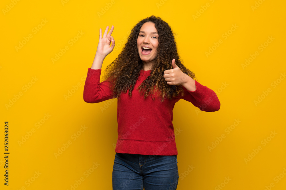 Teenager girl with red sweater over yellow wall showing ok sign with and giving a thumb up gesture Teenager girl with red sweater over yellow wall showing ok sign with and giving a thumb up gesture