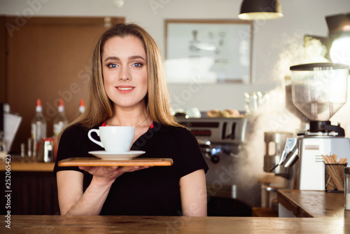 Beautiful female barista is working in coffee shop. Attractive woman is standing behind the bar counter, making coffee and welcomes customers.