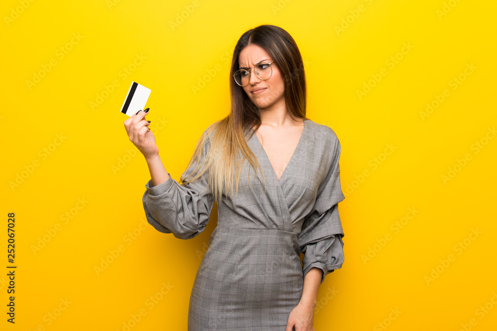 Young woman with glasses over yellow wall taking a credit card without money Young woman with glasses over yellow wall taking a credit card without money