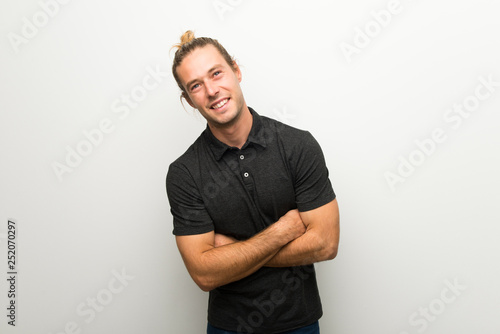 Blond man with long hair over white wall keeping the arms crossed while smiling