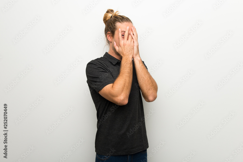 Blond man with long hair over white wall covering eyes by hands and looking through the fingers Blond man with long hair over white wall covering eyes by hands and looking through the fingers