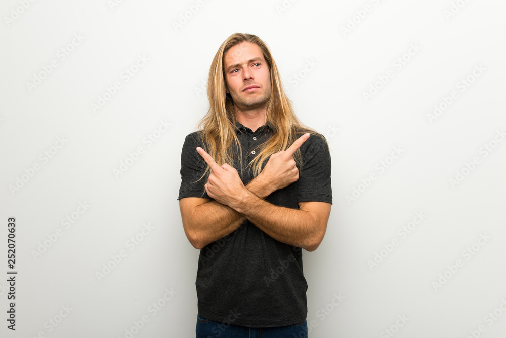 Blond man with long hair over white wall pointing to the laterals having doubts