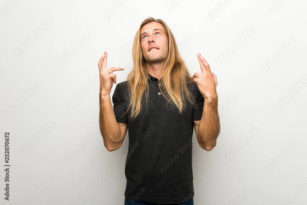 Blond man with long hair over white wall with fingers crossing and wishing the best Blond man with long hair over white wall with fingers crossing and wishing the best