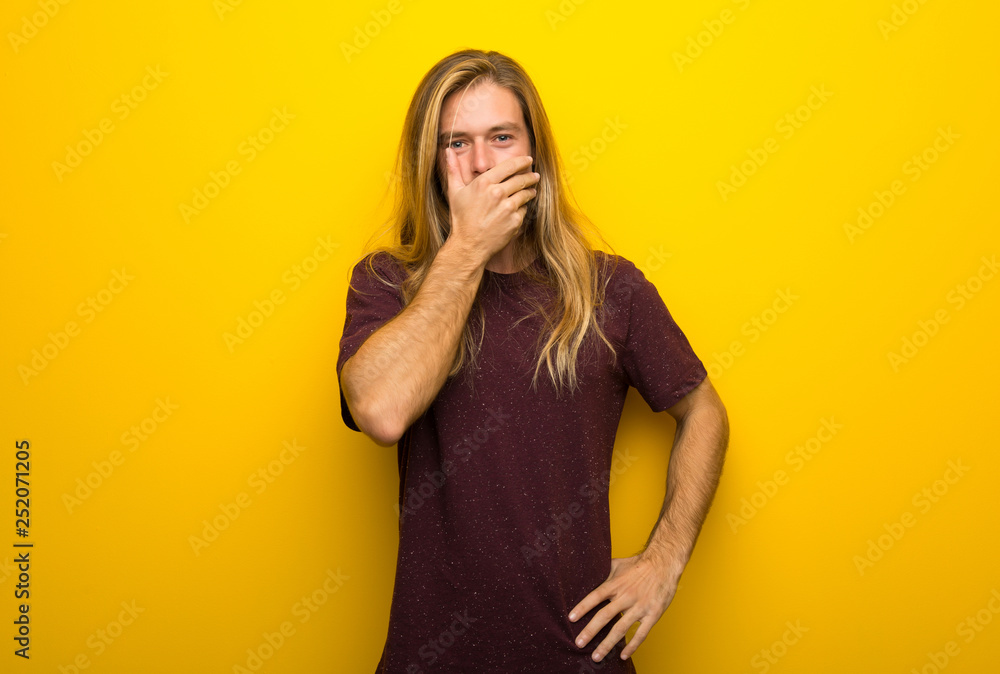 Blond man with long hair over yellow wall covering mouth with hands for saying something inappropriate Blond man with long hair over yellow wall covering mouth with hands for saying something inappropriate