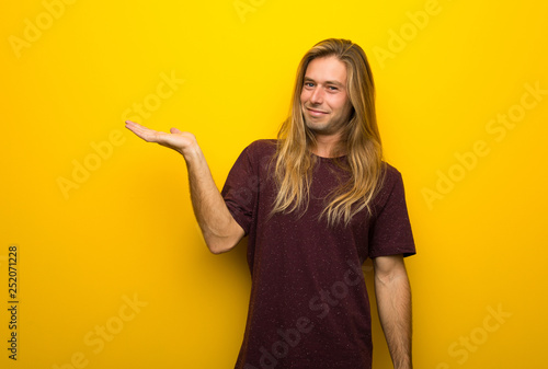 Blond man with long hair over yellow wall holding copyspace imaginary on the palm to insert an ad