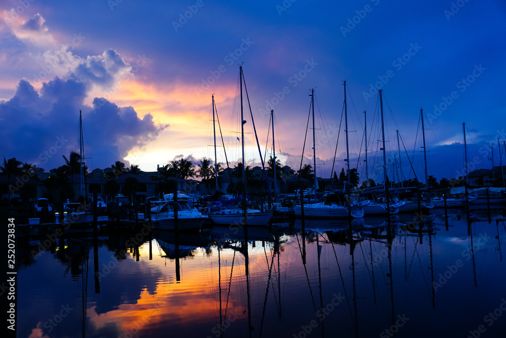 Sailboats reflecting on the water in a marina with blue and pink sunset and cloudy skies.