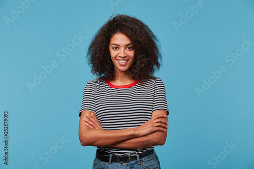 Self-confident happy young African American woman with long curly dark hair laughing smiling against blue background standing with arms crossed