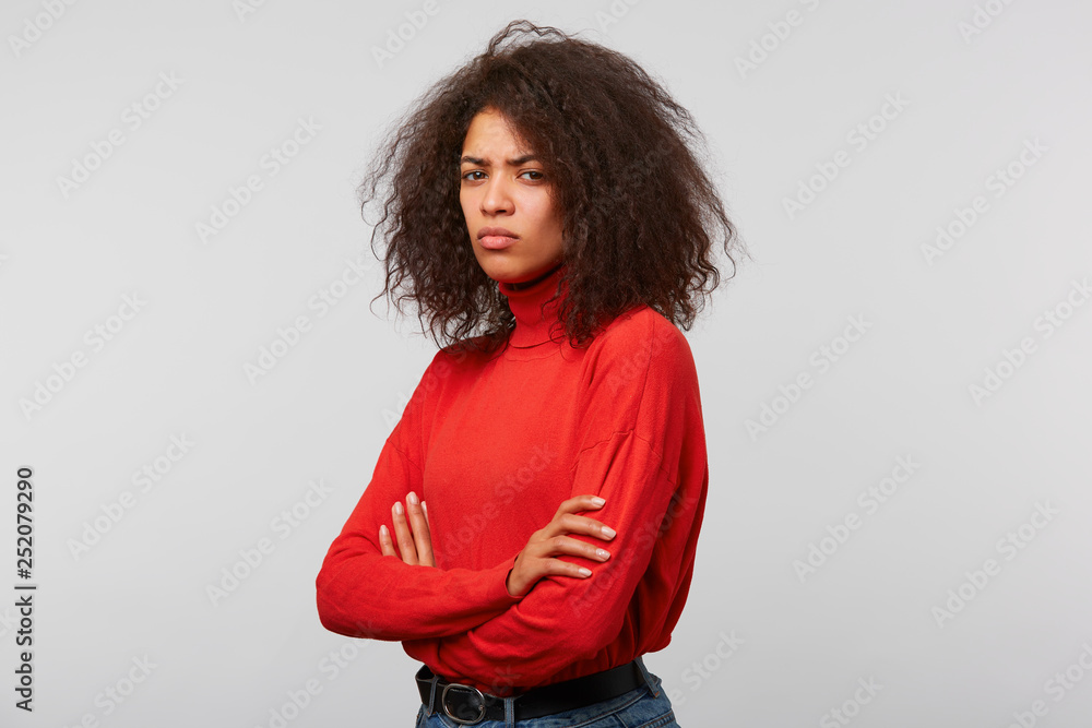 Studio photo of serious suspicious latino woman with curly afro hair standing half turned with crossed hands and frowning, expressing distrustfulness and disappointment, over white background - obrazy, fototapety, plakaty 