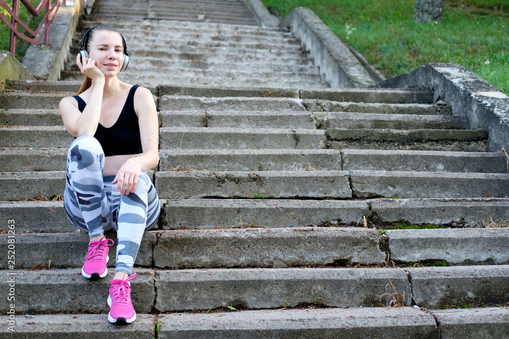 Young beautiful smiling slim sporty girl siting on the stairs and listen to music. Young beautiful smiling slim sporty girl siting on the stairs and listen to music.