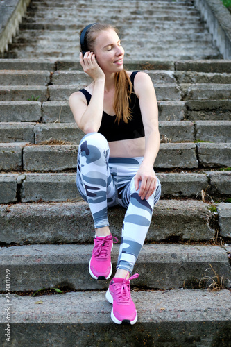 Young beautiful smiling slim sporty girl siting on the stairs and listen to music. Healthy sports lifestyle.