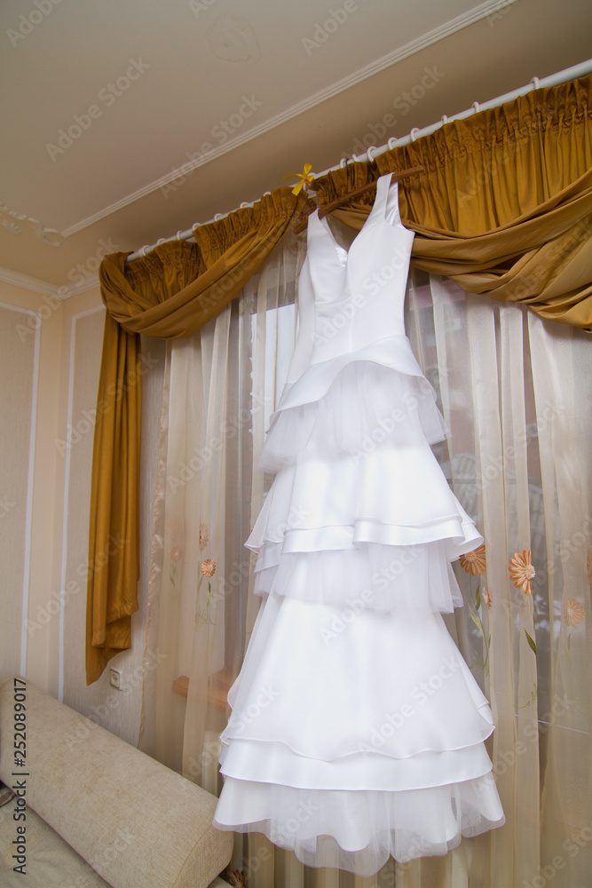 white bride dress hanging on a hanger in the room by the window without people white bride dress hanging on a hanger in the room by the window without people