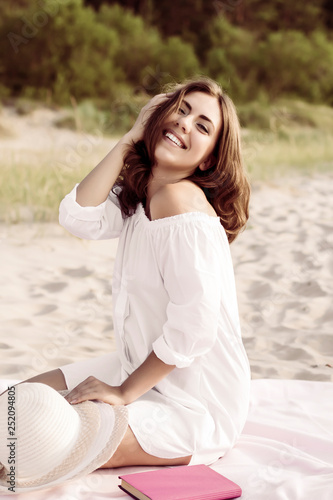 Young woman sitting at the beach wearing white straw hat. Happy girl enjoying summer vacation at the beach.