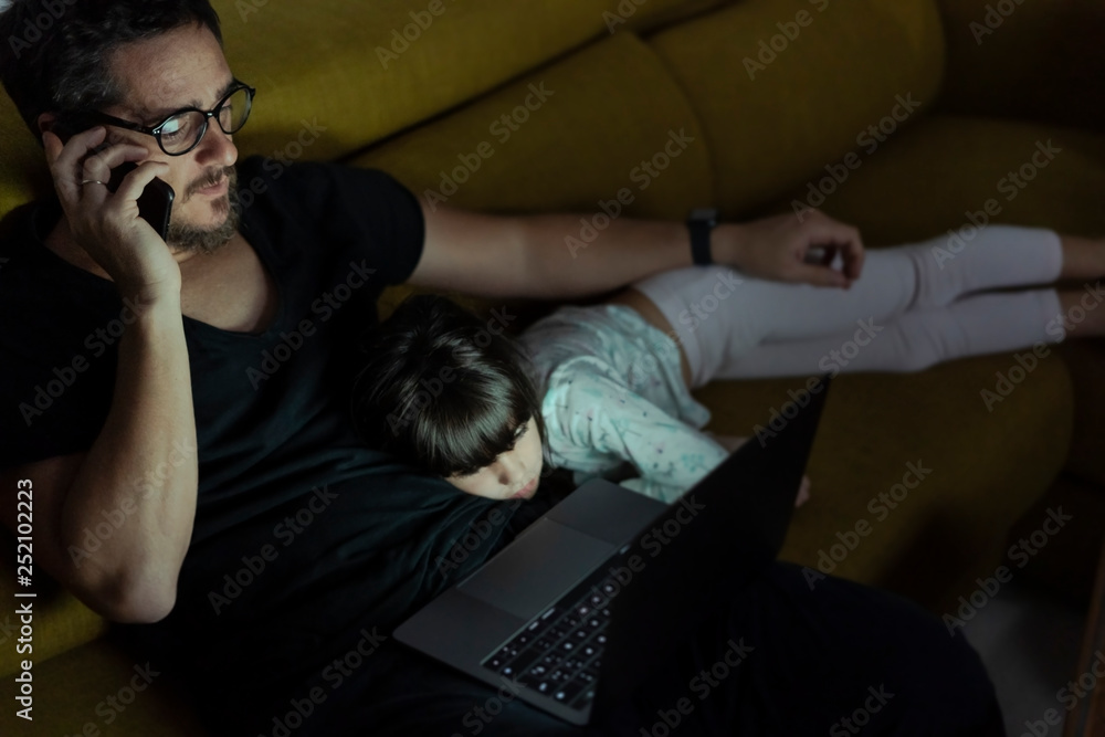 Father talking on cell phone on couch at night with daughter lying next to him Father talking on cell phone on couch at night with daughter lying next to him