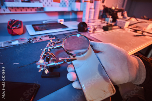 Electronic technician holds a modern smartphone with a broken body, carefully examines the damage using a magnifying glass sitting at the desk in the repair shop. Illumination with red and blue lights