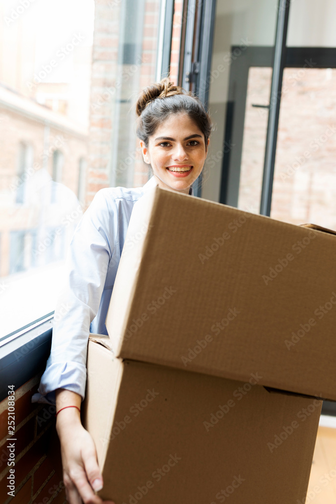 Portrait of smiling young woman carrying cardboard box in new apartment at the window