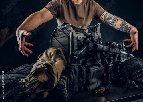 Casually dressed man showing his military uniform and equipment. Modern special forces equipment. Studio photo against a dark textured wall