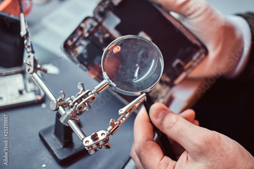 Repairman uses magnifier and tweezers to repair damaged smartphone. Close-up photo of a disassembled smartphone.