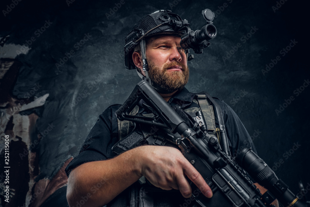 Fototapeta premium Bearded special forces soldier or private military contractor holding an assault rifle and observes the surroundings in night vision goggles. Studio photo against a dark textured wall