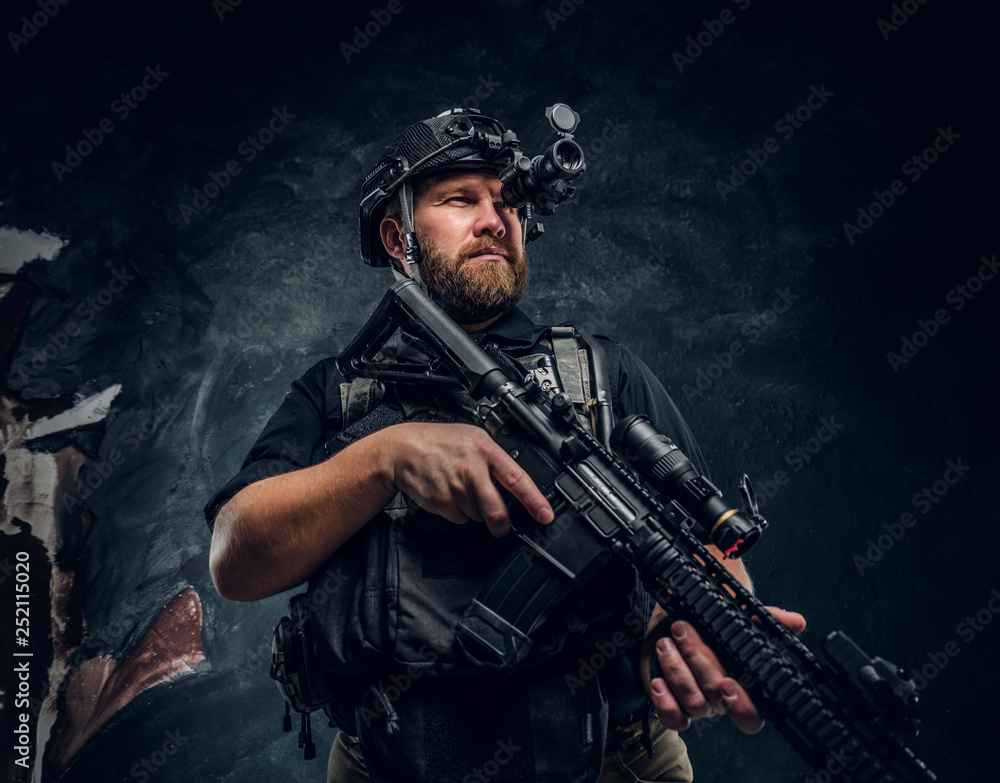 Fototapeta premium Bearded special forces soldier or private military contractor holding an assault rifle and observes the surroundings in night vision goggles. Studio photo against a dark textured wall