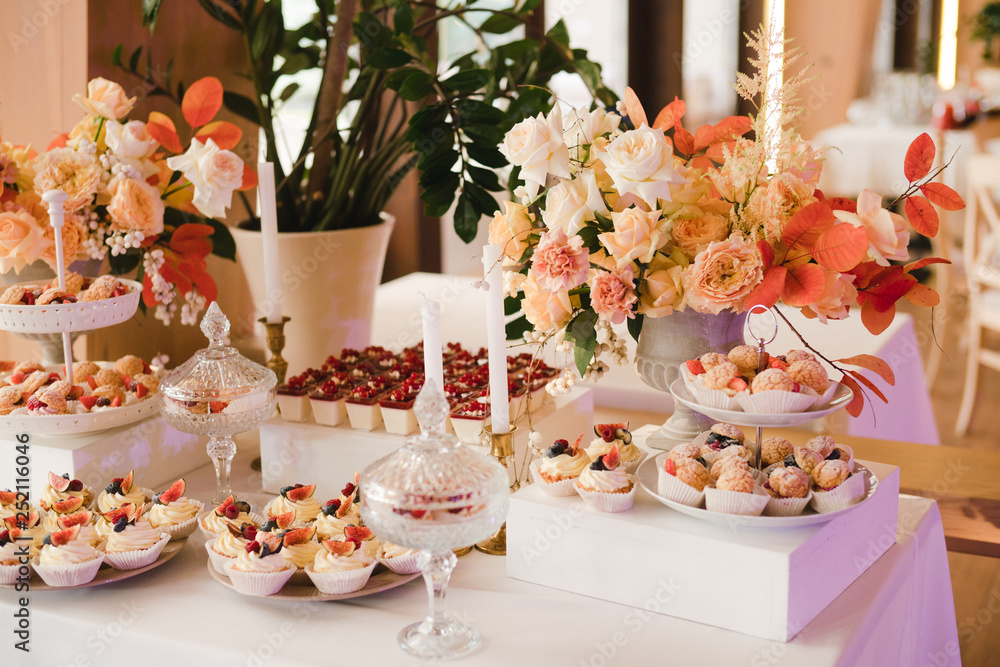 Candy bar with tasty desserts and appetizers at wedding reception. Decorated with flower compositions and candles. Sweet food, dessert, buffet, catering table, restaurant.